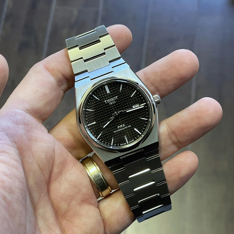 Stainless steel Tissot PRX watch with black textured dial and date display held in hand